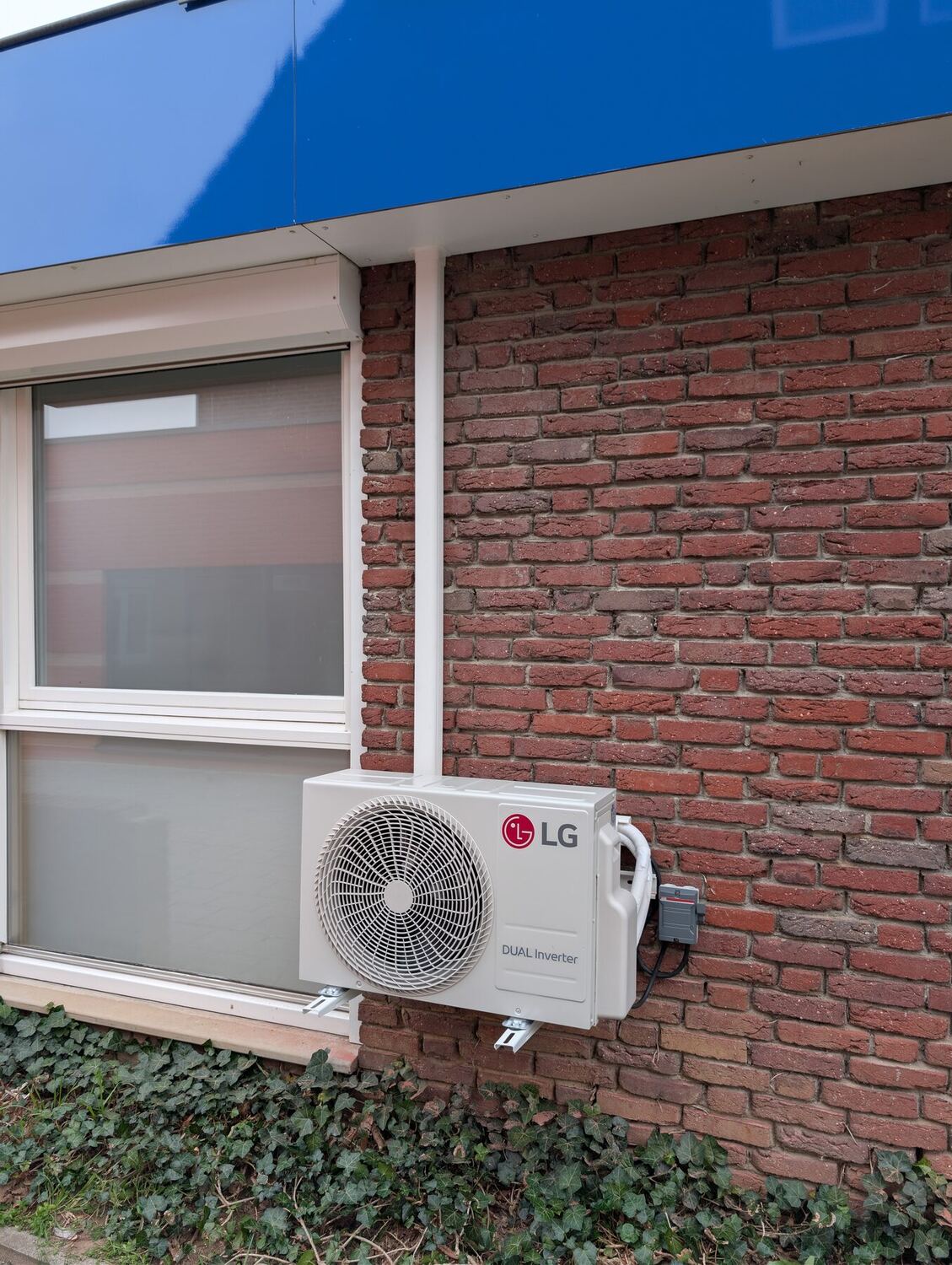 LG airco-buitenunit aan een bakstenen gevel onder een raam met rolluik
